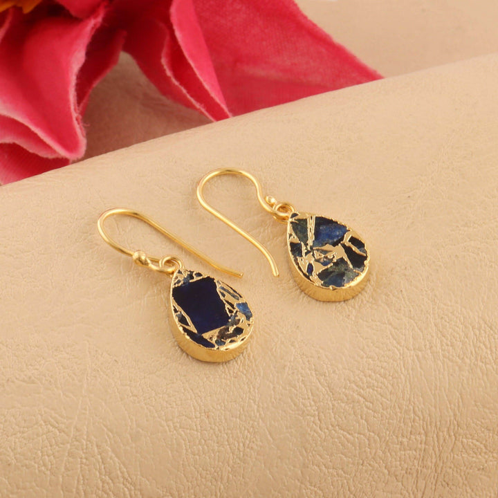 Gold earrings with blue stone drops on a beige surface with pink flowers in the background