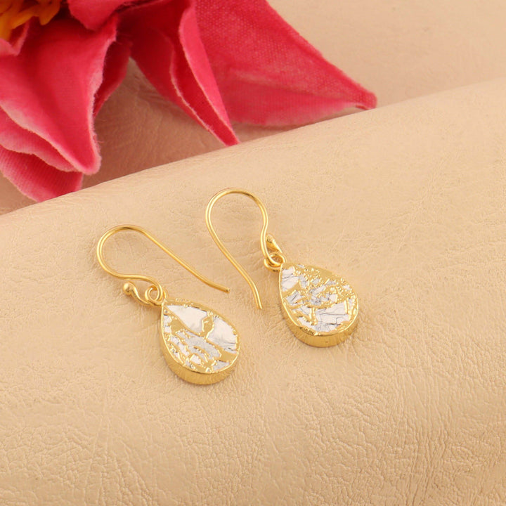 Gold earrings with clear stones on a beige surface with a pink flower in the background
