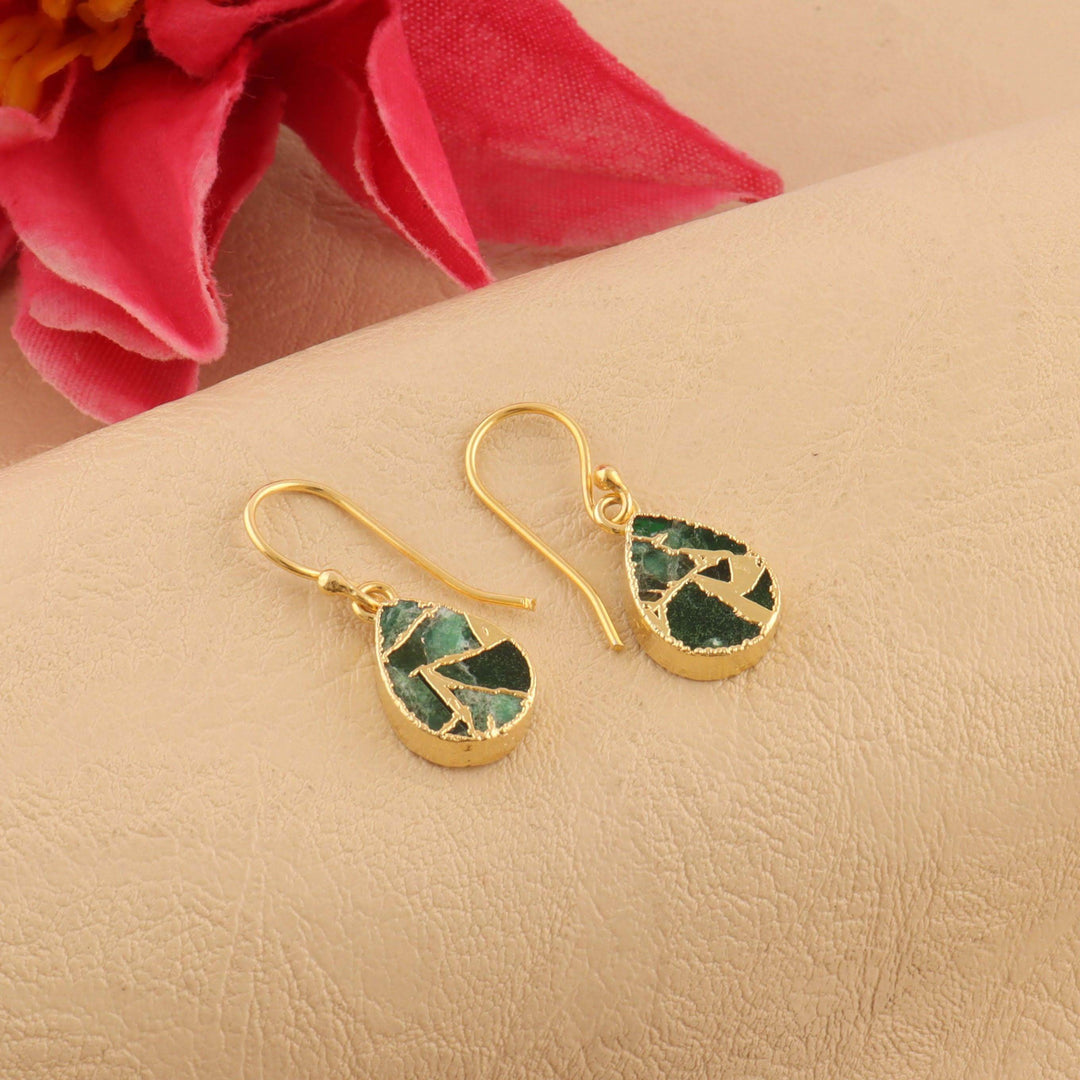 Gold earrings with green stone accents on a beige surface with pink fabric in the background