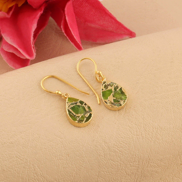 Gold earrings with green leaf-like stones on a beige surface with pink flowers in the background