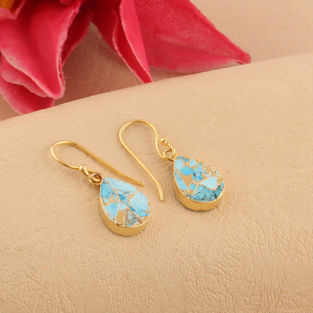 Gold earrings with turquoise stones on a beige surface with pink fabric in the background