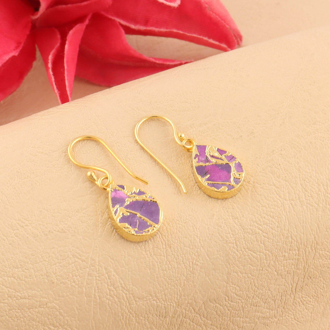 Gold earrings with purple gemstones on a beige surface with a red flower in the background