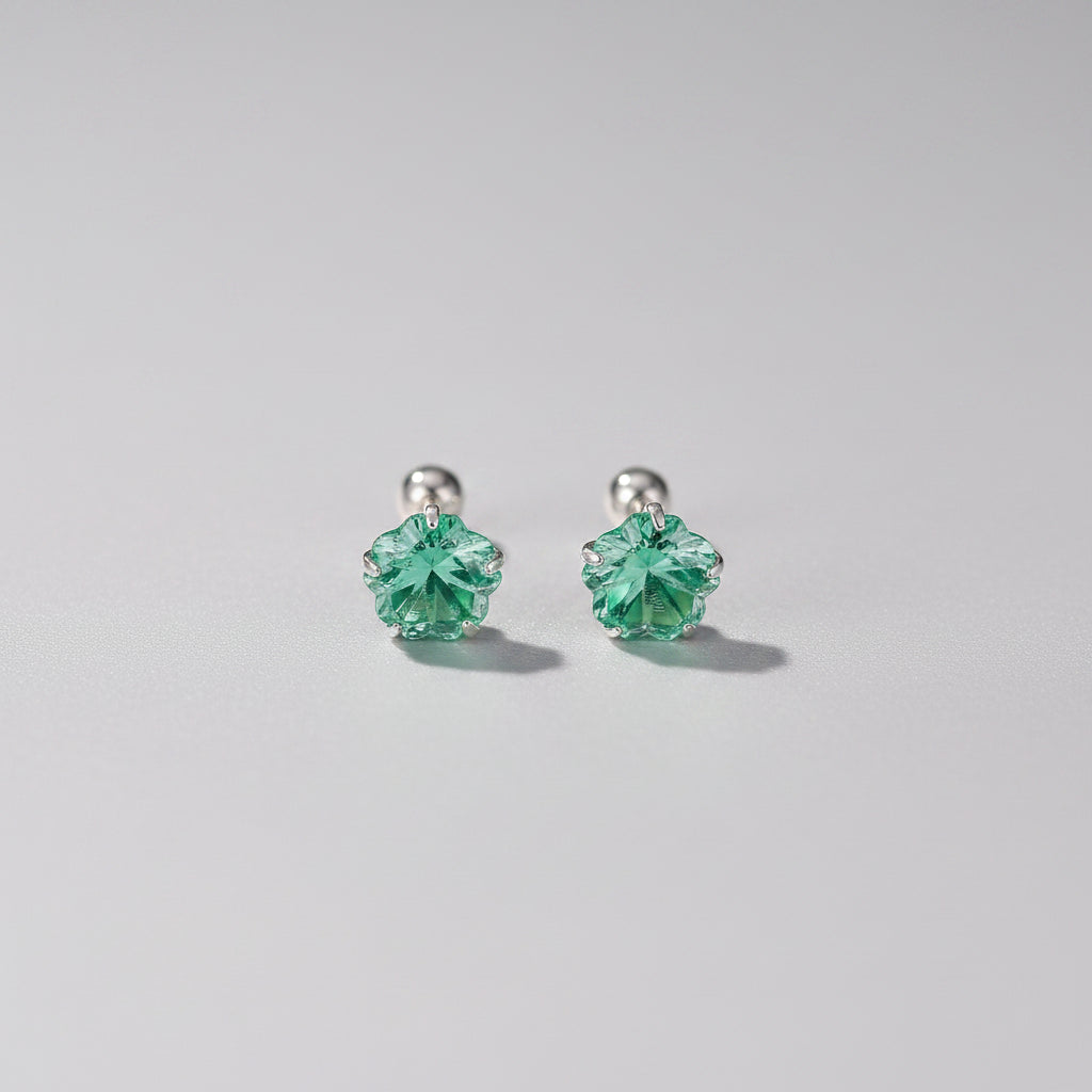A pair of silver 999 stud earrings with green faceted flower-shaped crystals, displayed on a light pink surface with decorative props.