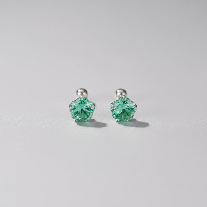 A pair of silver 999 stud earrings with green faceted flower-shaped crystals, displayed on a light pink surface with decorative props.