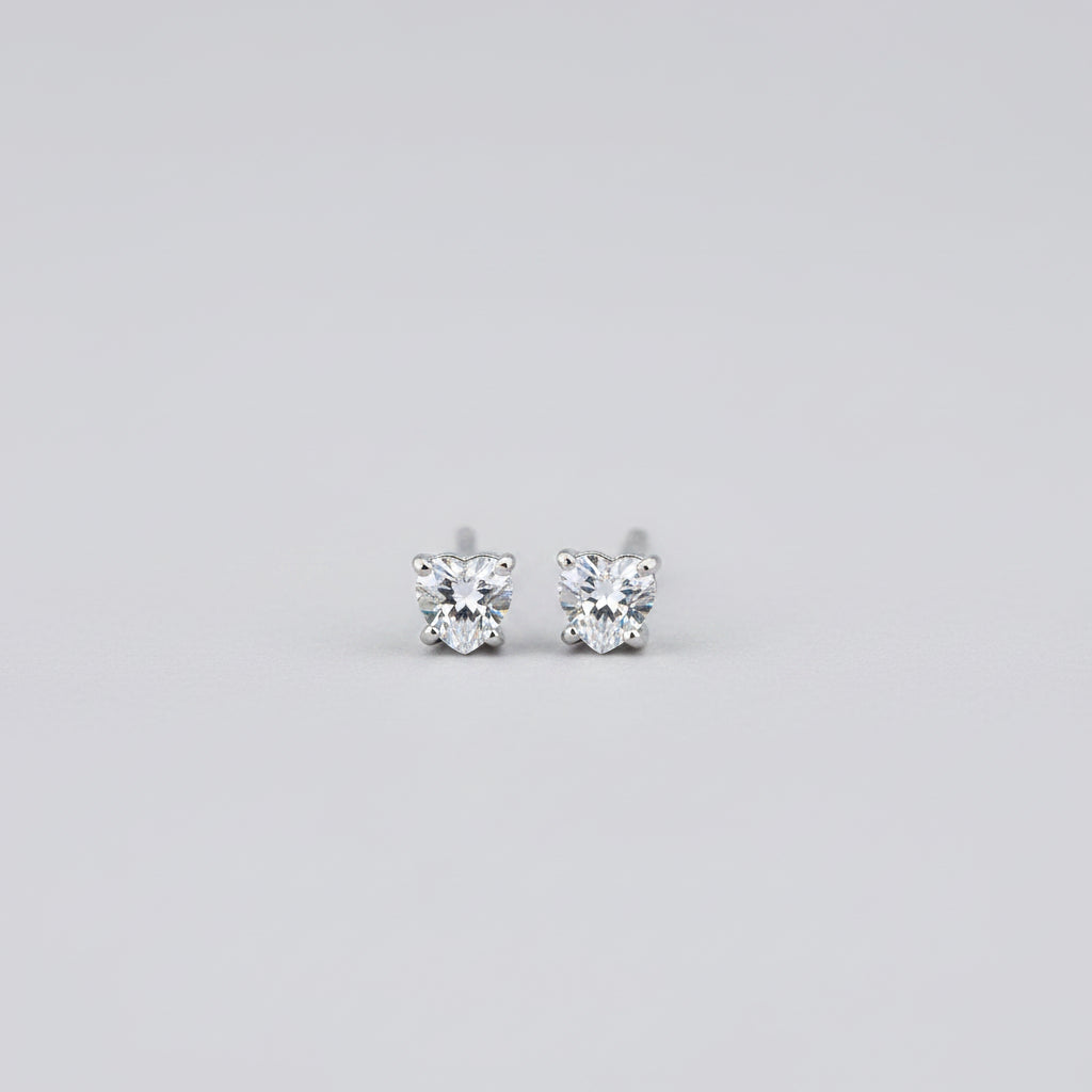 A close-up of a pair of small silver heart-shaped stud earrings with clear gemstones, set against a soft-focus jewelry box background.
