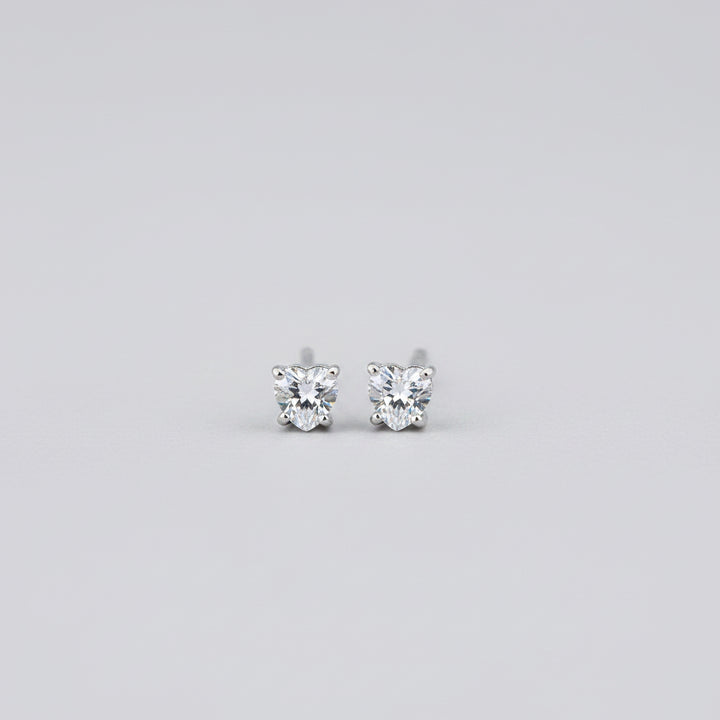 A close-up of a pair of small silver heart-shaped stud earrings with clear gemstones, set against a soft-focus jewelry box background.