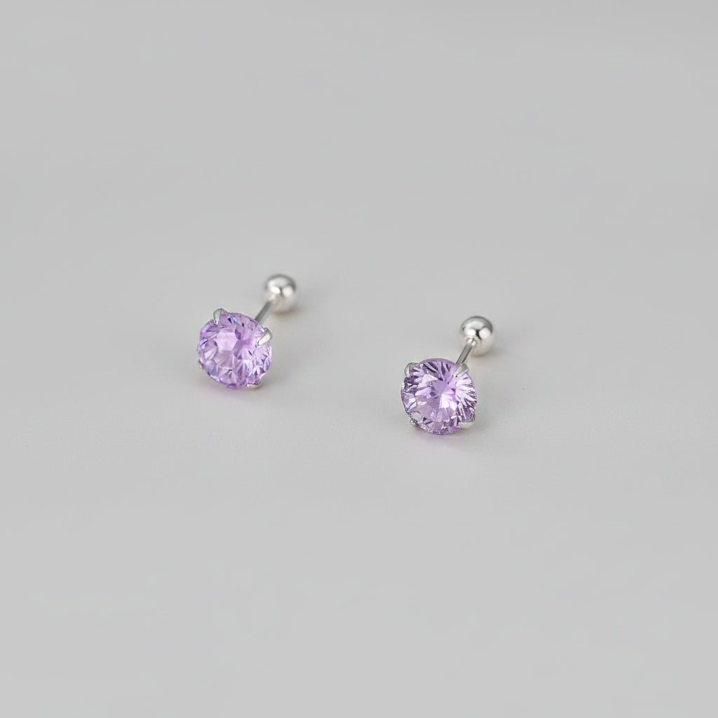 A pair of stud earrings with round faceted light purple gemstones in silver settings, displayed on a soft beige surface with blurred white flowers in the background. Text at the bottom reads &