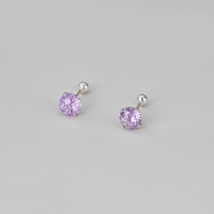 A pair of stud earrings with round faceted light purple gemstones in silver settings, displayed on a soft beige surface with blurred white flowers in the background. Text at the bottom reads &