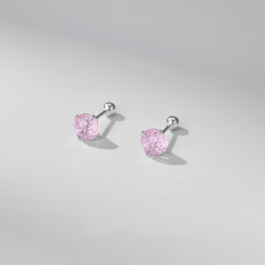 A pair of silver stud earrings with heart-shaped pink gemstones, displayed on a soft beige surface with decorative white flower petals. Text at the bottom reads &