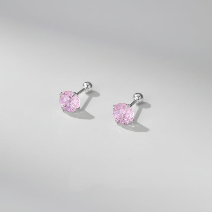 A pair of silver stud earrings with heart-shaped pink gemstones, displayed on a soft beige surface with decorative white flower petals. Text at the bottom reads &