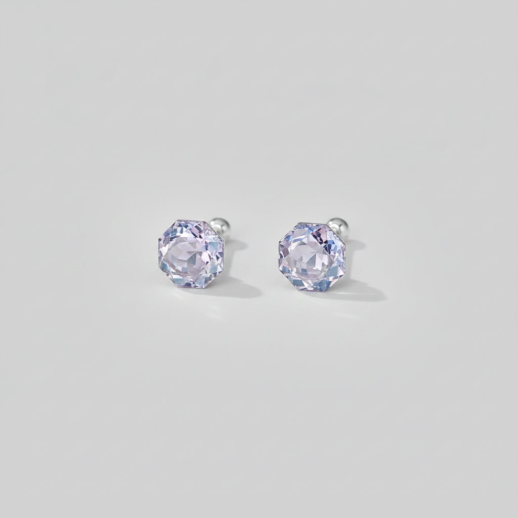 A pair of silver stud earrings with faceted light purple gemstones, displayed on a light beige surface with soft decorative shadows and petals.