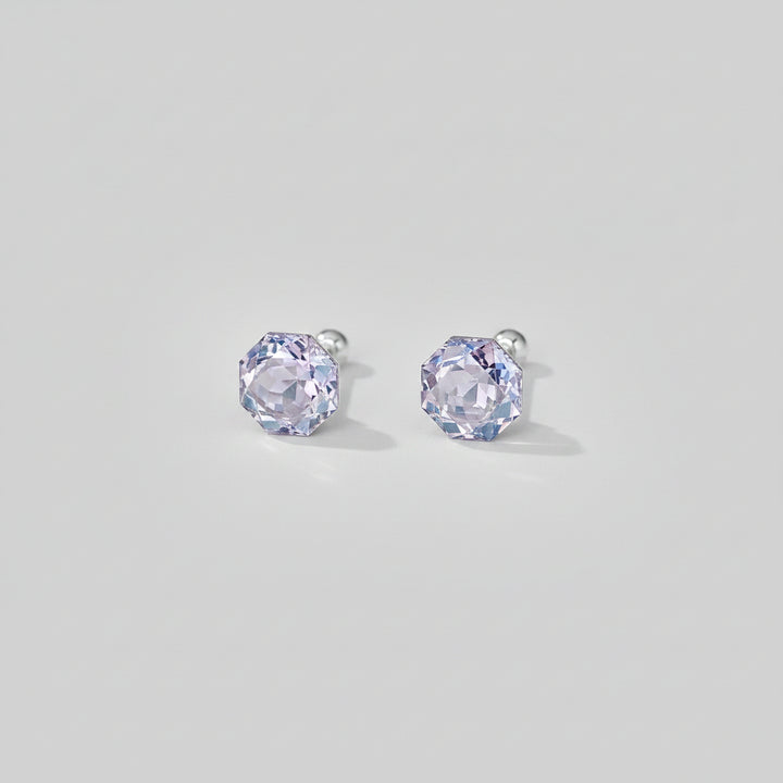 A pair of silver stud earrings with faceted light purple gemstones, displayed on a light beige surface with soft decorative shadows and petals.