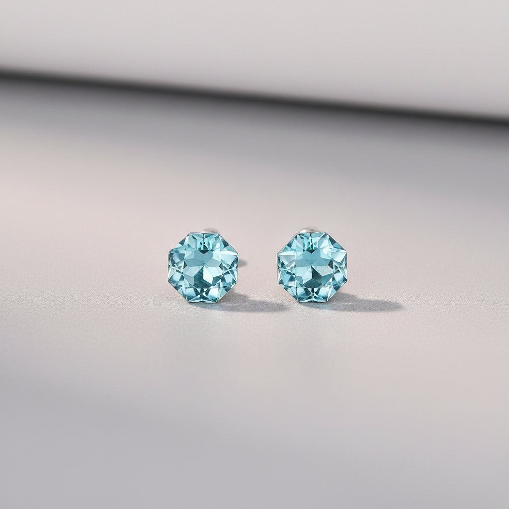 A pair of stud earrings with faceted light blue gemstones in silver settings, displayed on a light cream surface with soft decorative petals and a blurred orange object in the background. Text at the bottom reads &