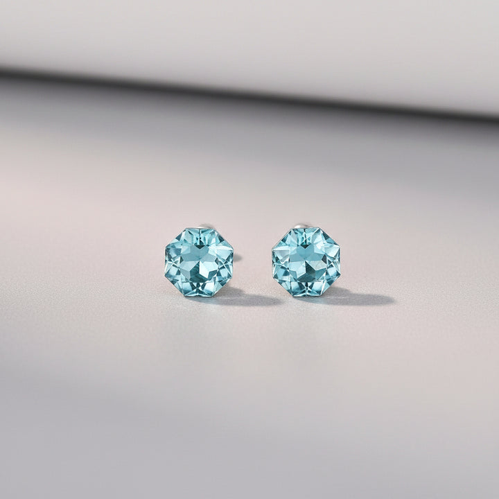 A pair of stud earrings with faceted light blue gemstones in silver settings, displayed on a light cream surface with soft decorative petals and a blurred orange object in the background. Text at the bottom reads &