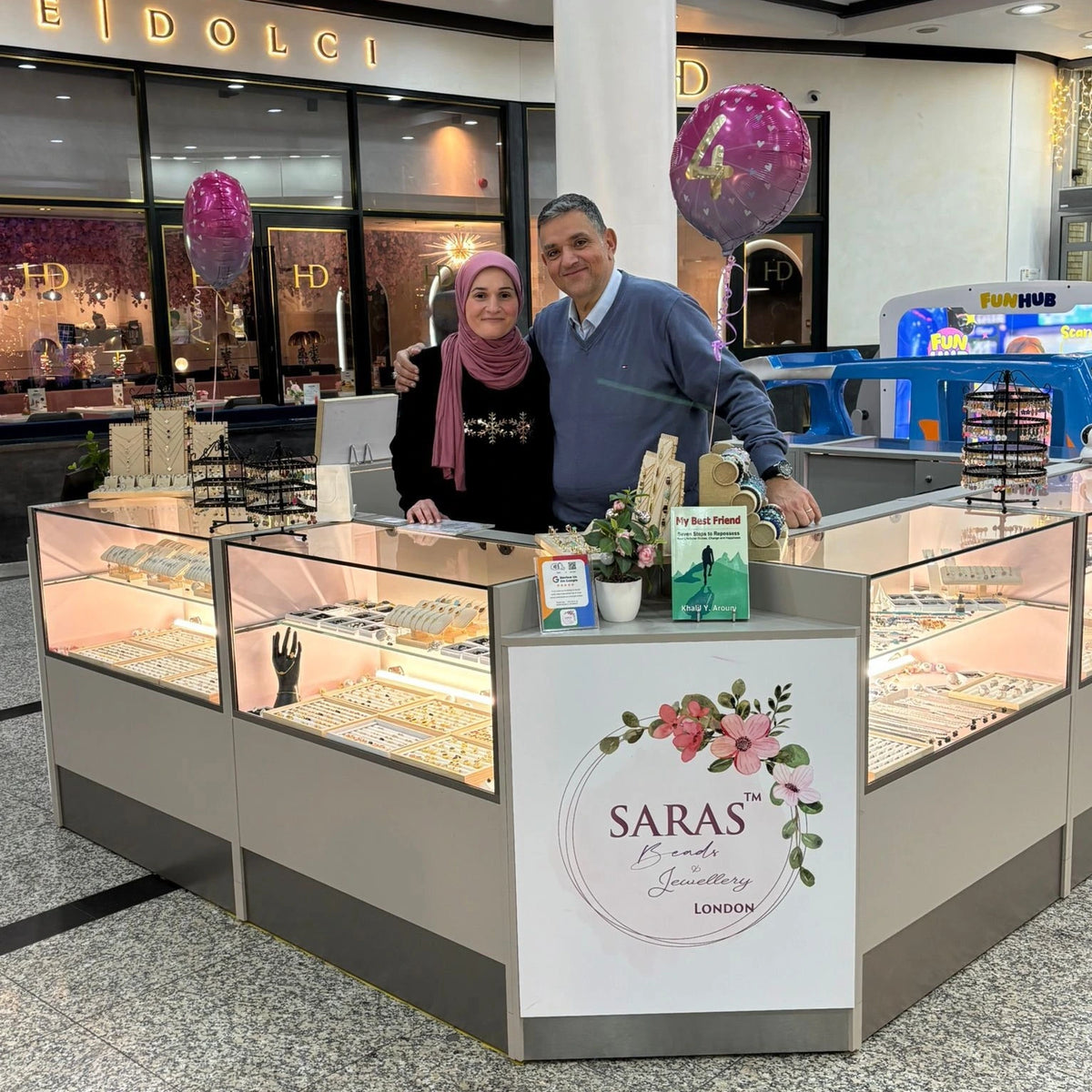 Two people standing behind a jewelry display counter with 'SARAS' branding in a shopping mall.